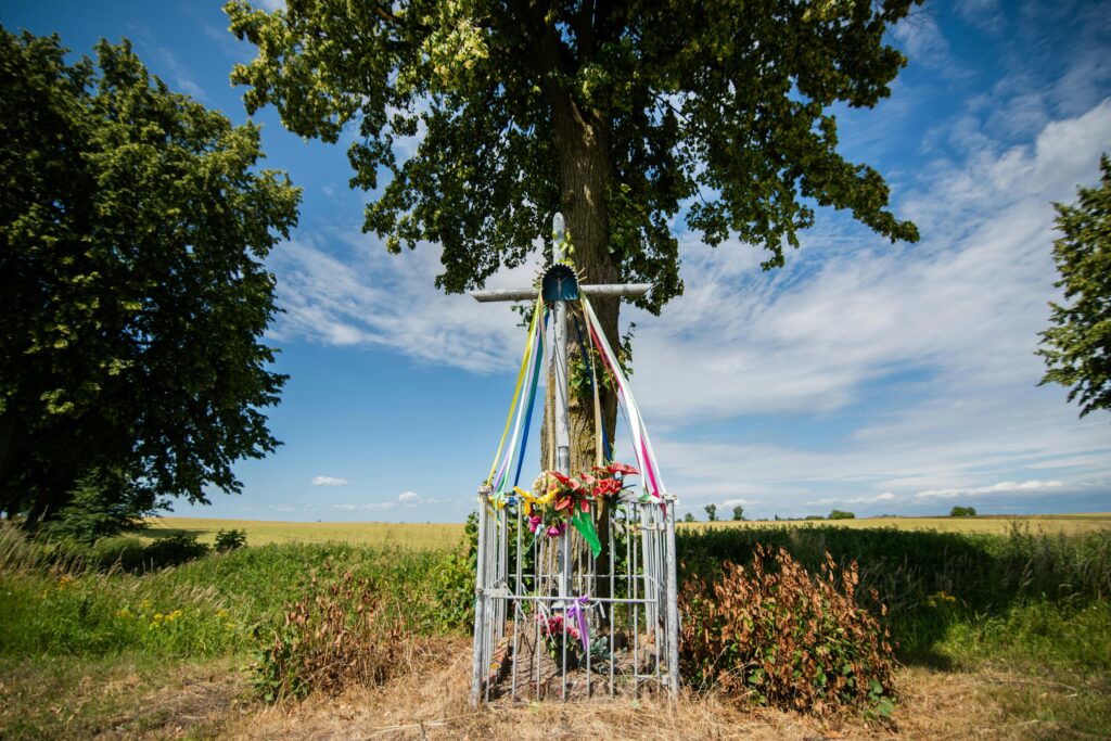 Scenic wayside shrine in Polish countryside, adorned with ribbons under blue sky.